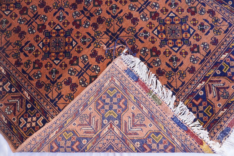 May include: An intricately patterned rug with a warm, reddish-orange background. The design features geometric shapes in navy blue, burgundy, and cream. The rug's corner is folded back, revealing a detailed underside with a similar color palette and fringe.