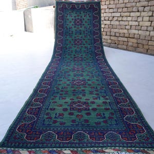 May include: A long, rectangular rug with a green field and intricate patterns. The rug features a dark blue border with floral and geometric designs in red and white. The rug is displayed on a light-colored surface.
