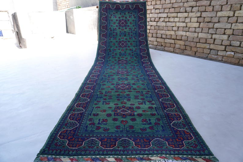 May include: A long, rectangular rug with a green field and intricate patterns. The rug features a dark blue border with floral and geometric designs in red and white. The rug is displayed on a light-colored surface.