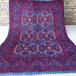 May include: A large, rectangular rug with a detailed, symmetrical pattern in shades of purple, blue, and red. The rug features a central design with repeating geometric and floral motifs, and a fringed edge. The rug is displayed on a white surface.