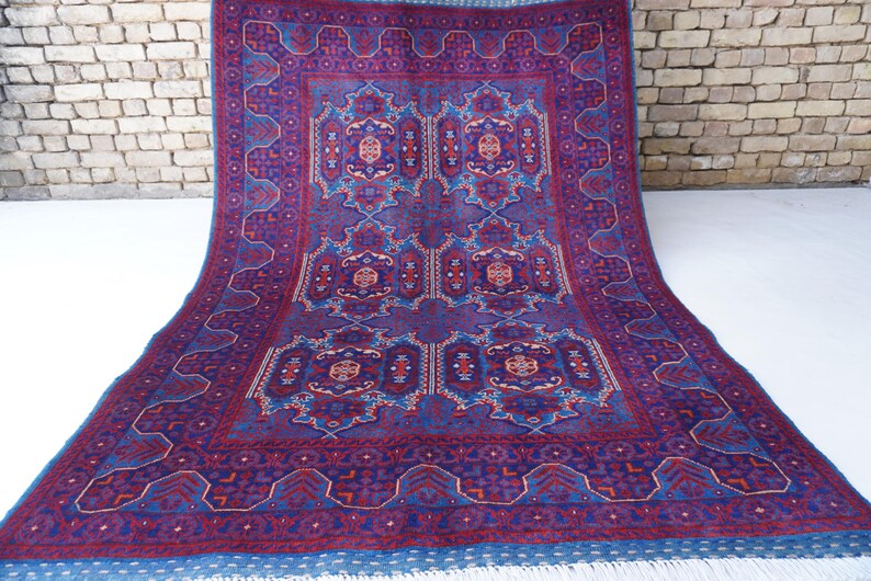 May include: A large, rectangular rug with a detailed, symmetrical pattern in shades of purple, blue, and red. The rug features a central design with repeating geometric and floral motifs, and a fringed edge. The rug is displayed on a white surface.