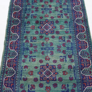 May include: A rectangular, hand-woven rug with a central field of teal green. The rug features a repeating pattern of floral and geometric designs in shades of blue, red, and white. The border has a detailed, stylized design.