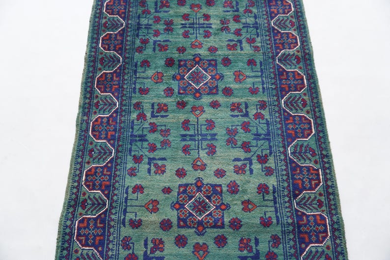 May include: A rectangular, hand-woven rug with a central field of teal green. The rug features a repeating pattern of floral and geometric designs in shades of blue, red, and white. The border has a detailed, stylized design.
