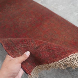 May include: A rectangular runner rug featuring a deep red base color with intricate, darker red and brown geometric patterns throughout. The rug has a short pile texture and is finished with light beige fringe along the short edge.