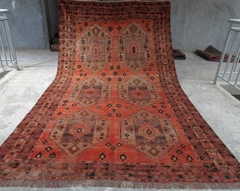 Puede incluir: Una alfombra rectangular grande que presenta un campo de color naranja vibrante con un patrón repetitivo de seis medallones geométricos simétricos en tonos marrón, gris y negro. La alfombra está enmarcada por un borde oscuro con motivos lineales.