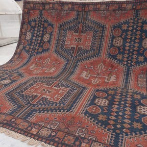 Pu&ograve; includere: Un grande tappeto rettangolare con un motivo geometrico nei toni blu navy, ruggine e beige. Il tappeto presenta un motivo a medaglione centrale e dettagli di bordo intricati. Il tappeto ha un bordo sfrangiato.