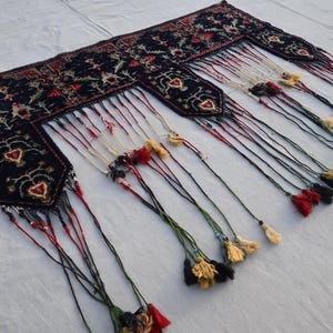 K&ouml;nnte beinhalten: Dunkelblaues, rechteckiges Textil mit aufwendigen Blumenmustern und herzf&ouml;rmigen Designs. Die R&auml;nder sind mit langen Quasten in verschiedenen Farben, darunter Rot, Schwarz und Gold, verziert. Es k&ouml;nnte sich um einen Teppich oder Wandbehang handeln.