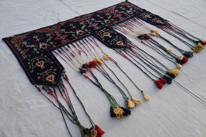 K&ouml;nnte beinhalten: Dunkelblaues, rechteckiges Textil mit aufwendigen Blumenmustern und herzf&ouml;rmigen Designs. Die R&auml;nder sind mit langen Quasten in verschiedenen Farben, darunter Rot, Schwarz und Gold, verziert. Es k&ouml;nnte sich um einen Teppich oder Wandbehang handeln.