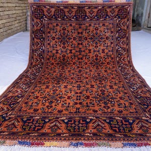 May include: An intricately patterned, rectangular rug with a warm orange field and a dark blue border. The rug features a repeating geometric design with accents of red, white, and blue. The rug has a fringed edge.
