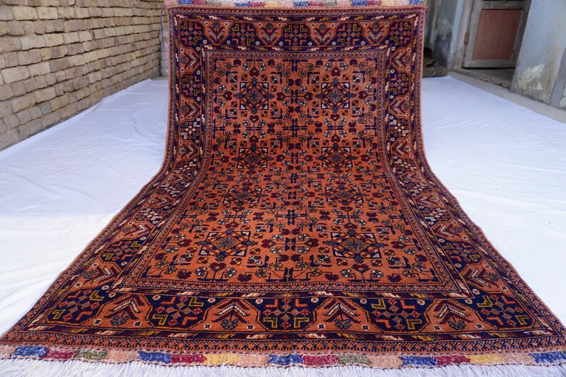 May include: An intricately patterned, rectangular rug with a warm orange field and a dark blue border. The rug features a repeating geometric design with accents of red, white, and blue. The rug has a fringed edge.