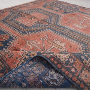 Pu&ograve; includere: Un tappeto antico con un motivo geometrico nei toni del rosso, blu e beige. Il tappeto presenta un design dettagliato con forme ripetute e un bordo piegato, che mostra il lato inferiore con un motivo blu.