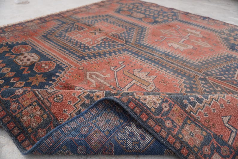 Pu&ograve; includere: Un tappeto antico con un motivo geometrico nei toni del rosso, blu e beige. Il tappeto presenta un design dettagliato con forme ripetute e un bordo piegato, che mostra il lato inferiore con un motivo blu.