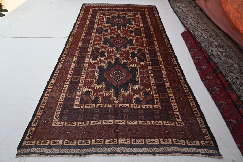 May include: A rectangular, hand-woven rug with a central geometric design in black, brown, and beige. The rug features a repeating border pattern and is displayed on a white surface. The rug's dimensions are not visible.