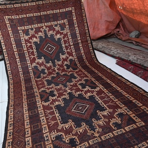 May include: A long, rectangular rug with a geometric pattern in shades of burgundy, brown, and navy blue. The rug features a central diamond design and intricate border details. Other rugs are stacked in the background.