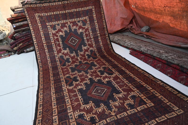 May include: A long, rectangular rug with a geometric pattern in shades of burgundy, brown, and navy blue. The rug features a central diamond design and intricate border details. Other rugs are stacked in the background.
