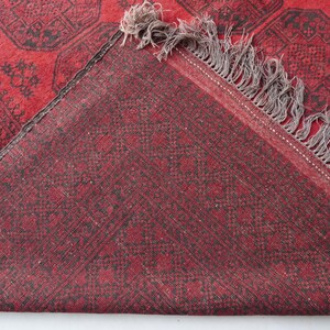 May include: A section of a red rug with a geometric pattern in black. The rug has a fringe on one edge and is folded over, revealing the underside, which is also red with a similar pattern. The rug appears to be made of woven material.