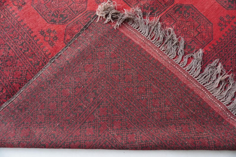 May include: A section of a red rug with a geometric pattern in black. The rug has a fringe on one edge and is folded over, revealing the underside, which is also red with a similar pattern. The rug appears to be made of woven material.