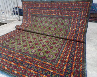 May include: A rectangular area rug featuring a central olive green field with a repeating pattern of dark red and white geometric floral motifs. The rug is bordered by multiple bands of intricate red, blue, and orange geometric designs.