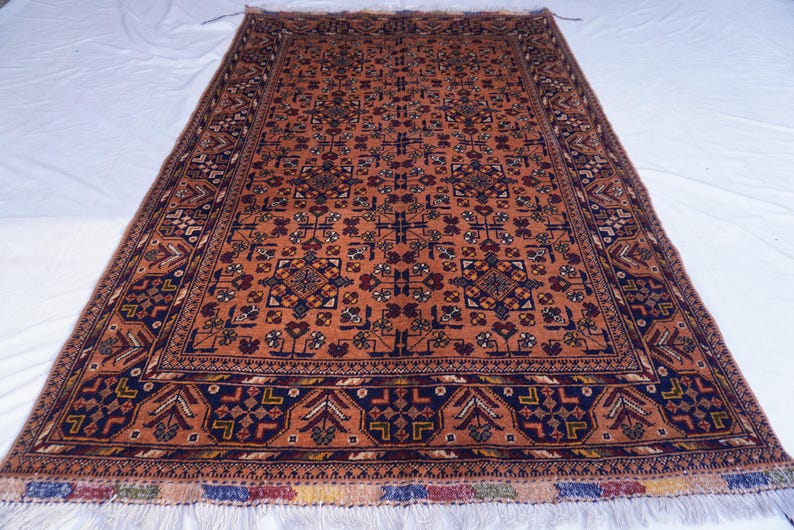 May include: An orange and navy rectangular rug with an intricate geometric pattern. The rug has a dark navy border with repeating shapes and a fringed edge. The central design features repeating floral and geometric motifs.