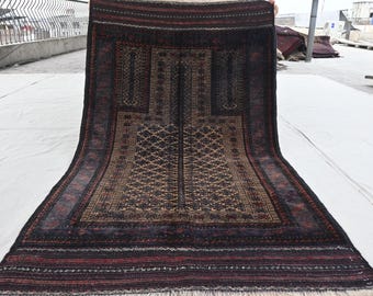May include: A rectangular, hand-woven rug with a central geometric design in shades of brown, black, and red. The rug features a detailed pattern with a border of stripes and fringe along the bottom edge. The rug is displayed outdoors.