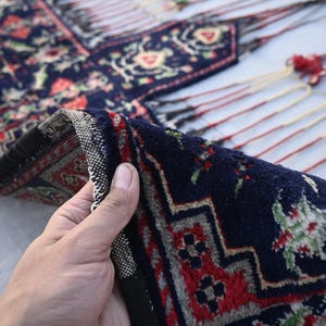 Puede incluir: Una secci&oacute;n de una alfombra azul oscuro con intrincados patrones florales y geom&eacute;tricos en rojo, blanco y verde. El borde de la alfombra est&aacute; doblado, revelando un borde detallado y flecos.