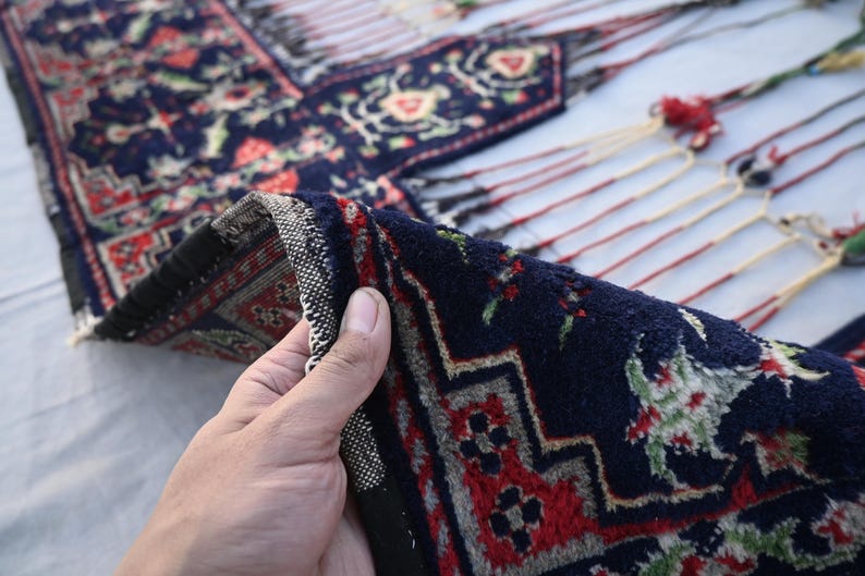 Puede incluir: Una secci&oacute;n de una alfombra azul oscuro con intrincados patrones florales y geom&eacute;tricos en rojo, blanco y verde. El borde de la alfombra est&aacute; doblado, revelando un borde detallado y flecos.