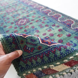 May include: A hand holds a section of a vintage runner rug. The rug features a teal background with a repeating floral pattern in red and blue. The border is dark green with a geometric design and fringe.
