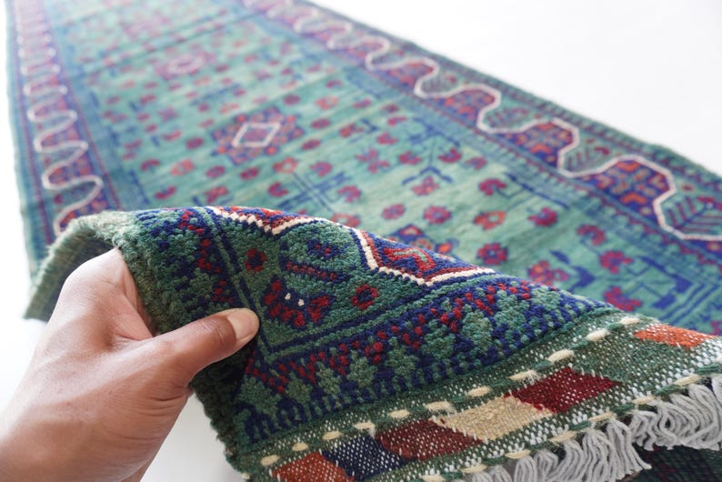 May include: A hand holds a section of a vintage runner rug. The rug features a teal background with a repeating floral pattern in red and blue. The border is dark green with a geometric design and fringe.
