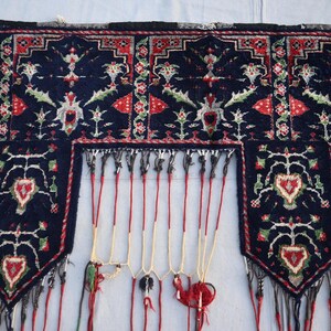 Puede incluir: Textil azul oscuro con intrincados patrones florales y geom&eacute;tricos en rojo, verde y blanco. El textil presenta un borde con flecos y cordones rojos colgantes, creando un elemento decorativo. El dise&ntilde;o incluye motivos en forma de coraz&oacute;n.