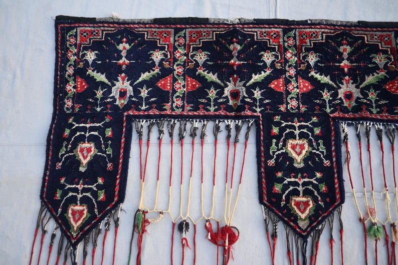 Puede incluir: Textil azul oscuro con intrincados patrones florales y geom&eacute;tricos en rojo, verde y blanco. El textil presenta un borde con flecos y cordones rojos colgantes, creando un elemento decorativo. El dise&ntilde;o incluye motivos en forma de coraz&oacute;n.