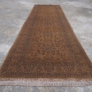 May include: A long, rectangular runner rug featuring a deep brown and tan color palette. The rug displays a detailed, symmetrical geometric pattern throughout the center and a wide, intricate border. It is finished with short, light-colored fringe.