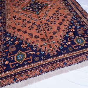 May include: An intricately patterned rug with a central diamond design and a border of repeating geometric shapes. The rug features a color palette of rust, navy blue, and touches of green and orange. The rug has a fringed edge.