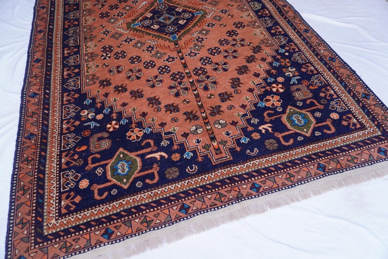 May include: An intricately patterned rug with a central diamond design and a border of repeating geometric shapes. The rug features a color palette of rust, navy blue, and touches of green and orange. The rug has a fringed edge.