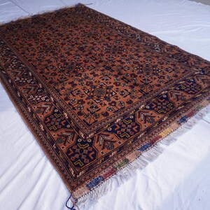 May include: An antique, rectangular rug with a detailed, geometric pattern in shades of rust, navy, and cream. The rug has a fringed edge and is displayed on a white surface. The rug's intricate design suggests a traditional, handcrafted origin.