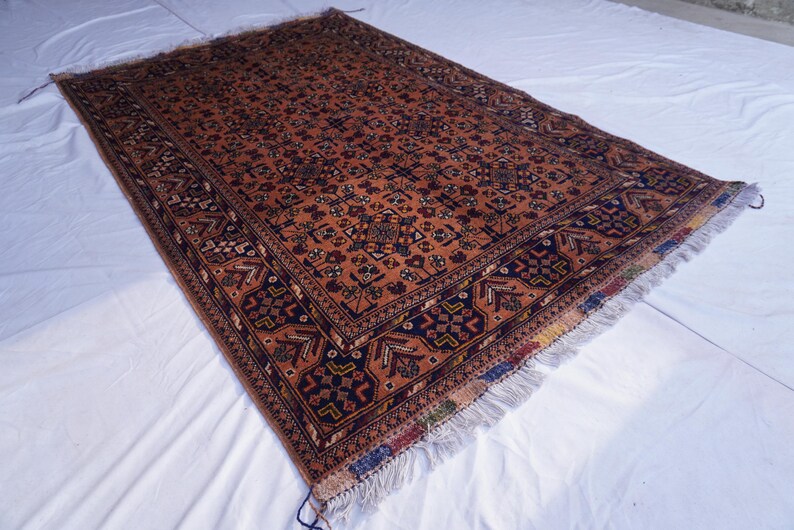 May include: An antique, rectangular rug with a detailed, geometric pattern in shades of rust, navy, and cream. The rug has a fringed edge and is displayed on a white surface. The rug's intricate design suggests a traditional, handcrafted origin.