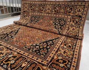 May include: A rectangular area rug featuring a complex pattern with a central rust-colored field and a wide black border. The design includes intricate floral and geometric motifs in shades of cream, gold, and brown, with the rug folded over.