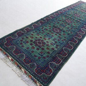 May include: A rectangular, hand-woven rug with a central field of green, adorned with geometric patterns in navy blue, red, and white. The rug features a detailed border and fringed edges, showcasing traditional craftsmanship. The rug's dimensions are not visible.