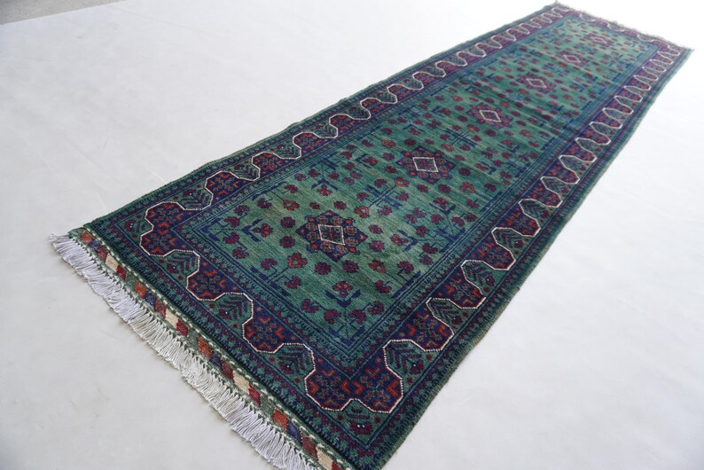 May include: A rectangular, hand-woven rug with a central field of green, adorned with geometric patterns in navy blue, red, and white. The rug features a detailed border and fringed edges, showcasing traditional craftsmanship. The rug's dimensions are not visible.