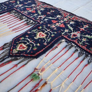 Puede incluir: Una alfombra azul marino con un detallado estampado floral en rojo, blanco y verde. La alfombra tiene un borde con flecos con borlas rojas, crema y negras. El dise&ntilde;o presenta flores y hojas estilizadas.