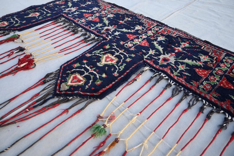 Puede incluir: Una alfombra azul marino con un detallado estampado floral en rojo, blanco y verde. La alfombra tiene un borde con flecos con borlas rojas, crema y negras. El dise&ntilde;o presenta flores y hojas estilizadas.