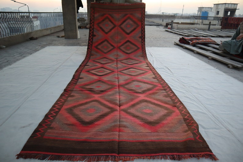 May include: A long, red and brown rug with a geometric diamond pattern. The rug is folded in half and laid out on a white surface.