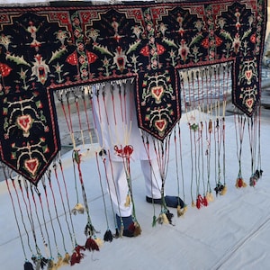 Puede incluir: Textil azul oscuro con patrones intrincados en rojo, verde y blanco, con dise&ntilde;os florales y geom&eacute;tricos. Tiene un borde con flecos y borlas multicolores. La pieza se sostiene, mostrando su detallada artesan&iacute;a.
