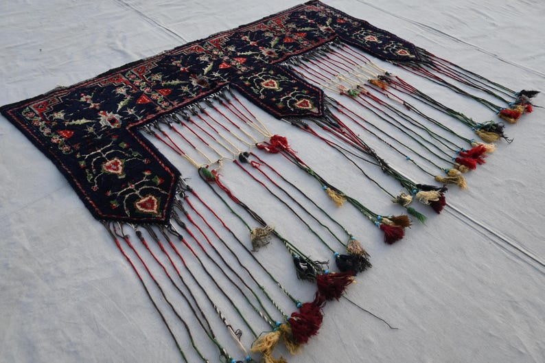 Puede incluir: Textil azul oscuro adornado con intrincados patrones florales y geom&eacute;tricos. El textil presenta m&uacute;ltiples secciones con largas borlas decorativas en varios colores, incluyendo rojo, blanco y verde. El dise&ntilde;o es sim&eacute;trico y detallado.