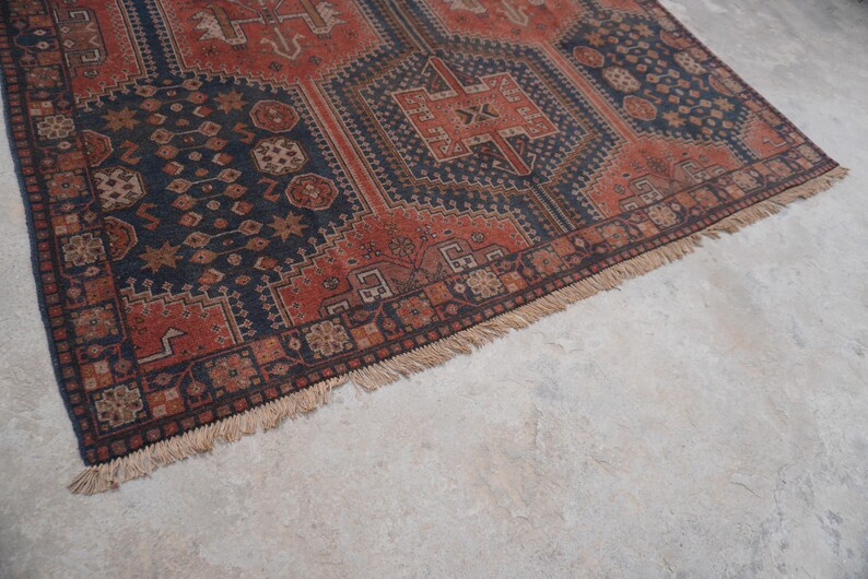 Pu&ograve; includere: Una porzione di un tappeto vintage con un motivo geometrico nei toni blu navy, ruggine e marrone. Il tappeto ha un bordo con frange ed &egrave; posto su una superficie grigio chiaro.