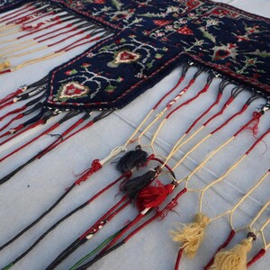 K&ouml;nnte beinhalten: Ein Abschnitt eines dunkelblauen Teppichs mit einem detaillierten Blumenmuster in Rot, Wei&szlig; und Gr&uuml;n. Der Teppich hat einen Fransenrand mit roten, schwarzen und cremefarbenen Quasten, die dem Textil ein dekoratives Element verleihen.