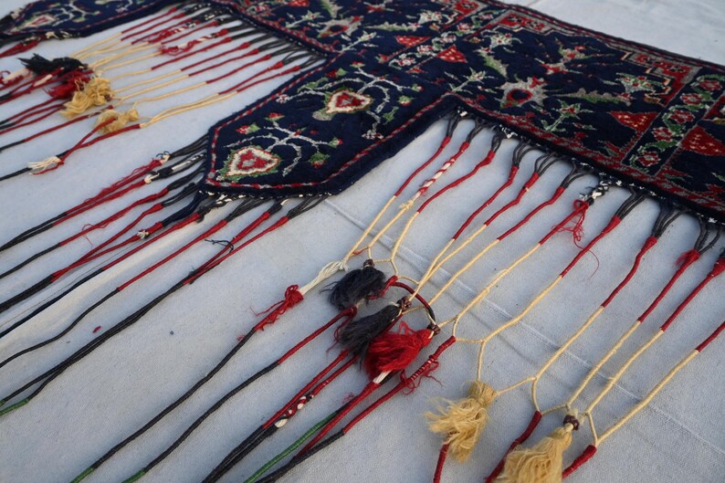 K&ouml;nnte beinhalten: Ein Abschnitt eines dunkelblauen Teppichs mit einem detaillierten Blumenmuster in Rot, Wei&szlig; und Gr&uuml;n. Der Teppich hat einen Fransenrand mit roten, schwarzen und cremefarbenen Quasten, die dem Textil ein dekoratives Element verleihen.