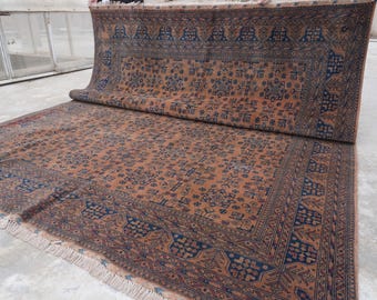 Könnte beinhalten: Ein großer, rechteckiger Teppich mit einem detaillierten, geometrischen Muster in Braun- und Marineblautönen. Der Teppich ist teilweise aufgerollt und zeigt sein kompliziertes Design und die gefransten Kanten. Der Teppich ist für die Inneneinrichtung geeignet.