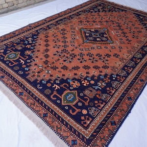 May include: Rectangular rug with a central terracotta field and intricate navy blue borders. The rug features geometric and floral patterns in shades of blue, green, and orange. The edges have a fringed finish.