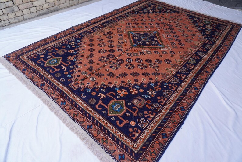 May include: Rectangular rug with a central terracotta field and intricate navy blue borders. The rug features geometric and floral patterns in shades of blue, green, and orange. The edges have a fringed finish.