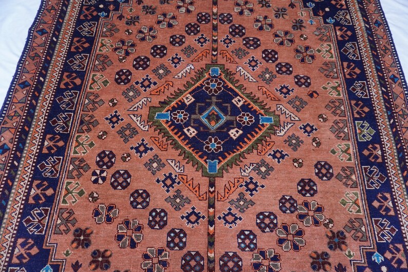 May include: A detailed, rectangular rug with a central diamond pattern in navy blue, green, and orange. The rug features a salmon-colored field with repeating floral and geometric motifs, and a navy blue border.